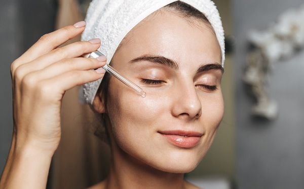 Người phụ nữ đội khăn trắng đang dùng ống nhỏ giọt thoa serum lên mặt, mắt nhắm, môi mỉm cười nhẹ.