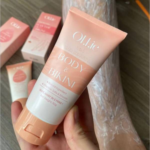 tay_sach_long_voi_kem_tay_ollie_50043f36a6494d24b721417dee714002_grande Tuýp kem tẩy lông màu hồng cầm trên tay, nền có hộp và tuýp nhỏ hơn.