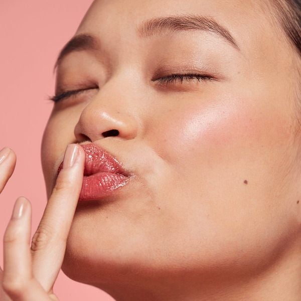 Người với l&agrave;n da s&aacute;ng, mắt nhắm, đang chạm ng&oacute;n tay l&ecirc;n đ&ocirc;i m&ocirc;i hồng, nền m&agrave;u hồng nhạt.