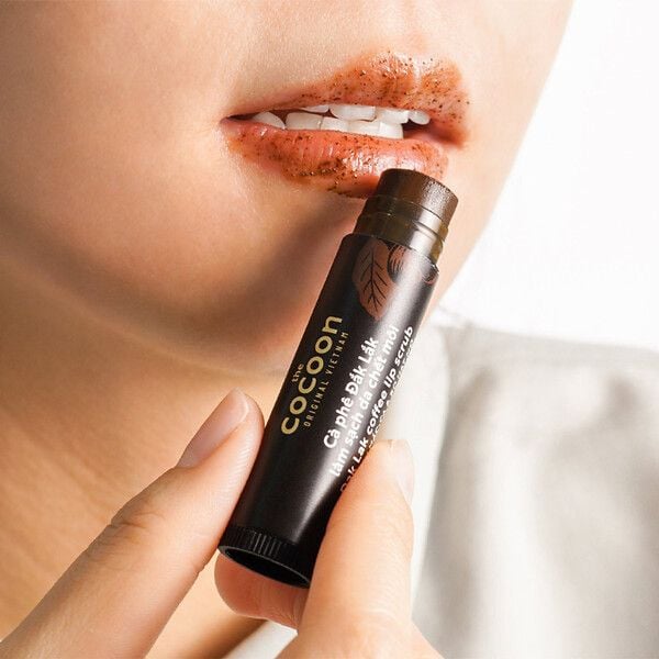 Người cầm thỏi tẩy tế b&agrave;o chết m&ocirc;i gần miệng, m&ocirc;i c&oacute; lớp tẩy tế b&agrave;o chết dạng hạt m&agrave;u n&acirc;u.