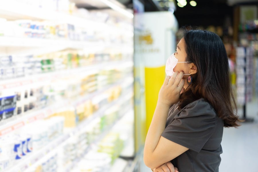Người phụ nữ đeo khẩu trang đang đứng trước kệ hàng trong siêu thị, nhìn vào các sản phẩm được trưng bày.
