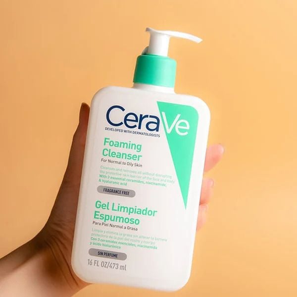 Chai sữa rửa mặt CeraVe Foaming Cleanser trên tay, nền màu cam nhạt.