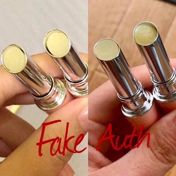 thoi_son_dhc_that_co_chat_son_ben_trong_mau_vang_nhat__khong_mui_7057fffb7a6f41bcad44cfa63526f3d9_grande So sánh hai thỏi son, một bên nhãn "Fake", bên kia nhãn "Auth", cầm trên tay, nền màu trung tính.