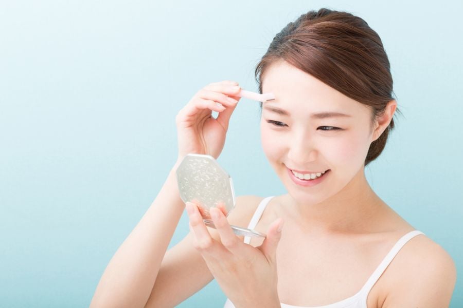 tia-long-may-nhi-4_b619f817d8734fb1b09641fe50a12e7c Người phụ nữ cầm gương nhỏ, dùng dụng cụ trang điểm lông mày, nền xanh nhạt.