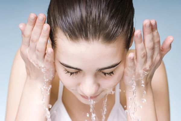 Người đang rửa mặt với nước, hai tay nâng nước lên, mắt nhắm và tóc ướt, nền xanh nhạt.