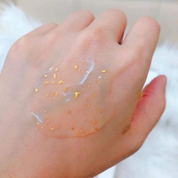 tinh_chat_vang_24k_la_gi_9ee999fb2bb54f79801e78fe8e894468_grande Gel trong suốt với hạt vàng nhỏ được thoa trên mu bàn tay, nền trắng mờ.