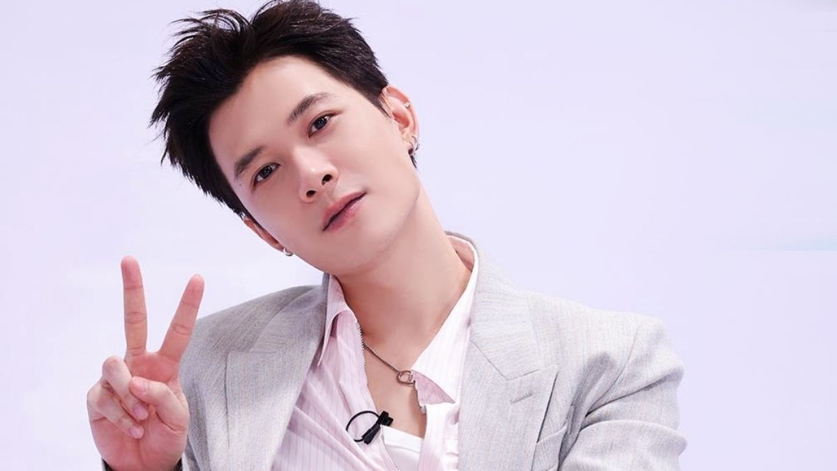 Người đàn ông mặc vest xám, áo sơ mi trắng, giơ hai ngón tay tạo dáng ký hiệu hòa bình, nền màu nhạt.