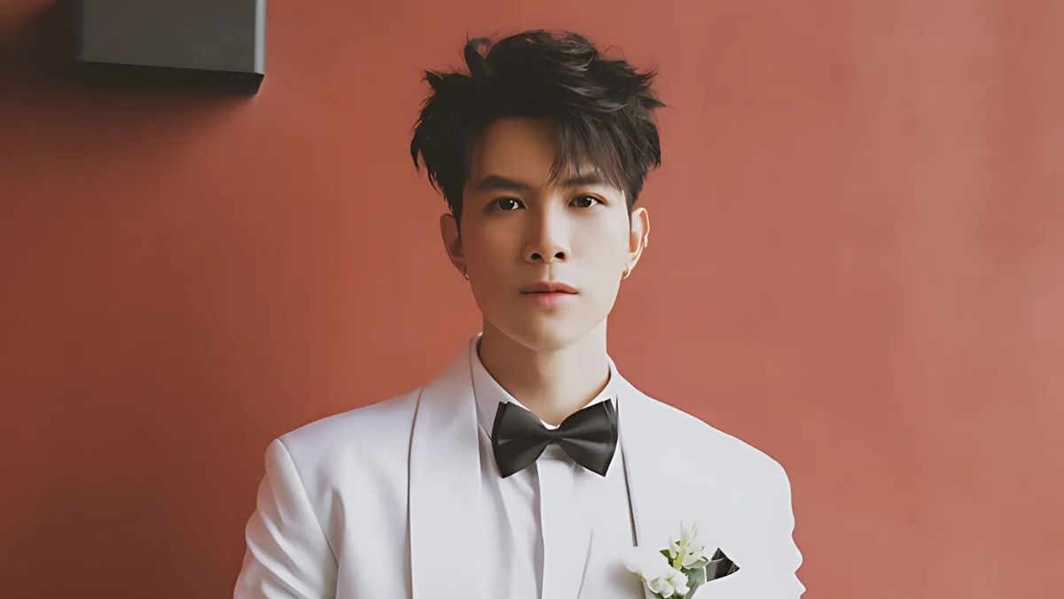 Người đàn ông mặc vest trắng, thắt nơ đen, đứng trước nền đỏ, cài hoa trắng ở áo.