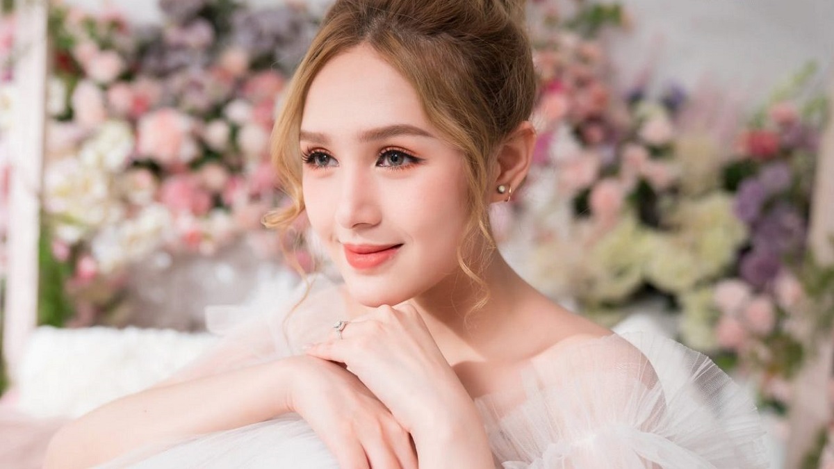 Người phụ nữ trẻ mỉm cười, mặc váy trắng, nền hoa mờ phía sau, ánh sáng dịu nhẹ.