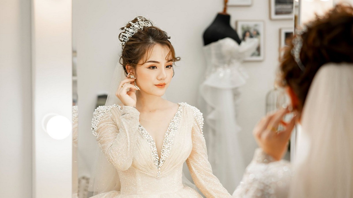 toc-co-dau-23 Người phụ nữ mặc váy cưới trắng, đội vương miện, chỉnh trang trước gương trong phòng trang điểm.