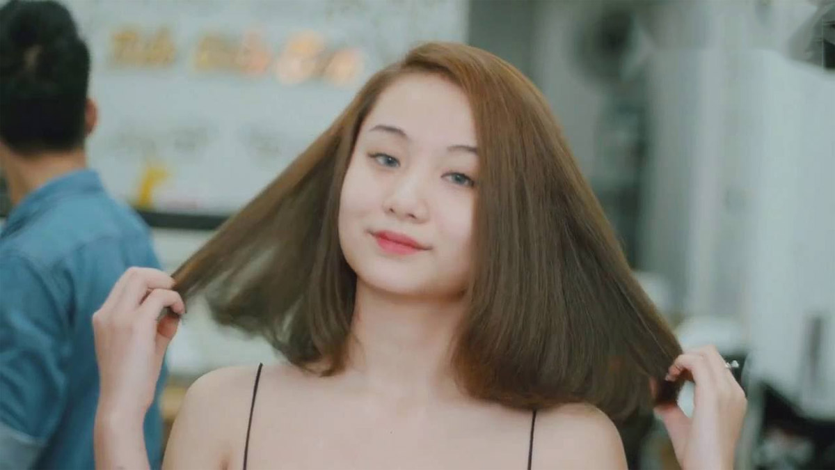 Người phụ nữ mỉm cười, đang vuốt tóc ngang vai, nền là tiệm làm tóc.