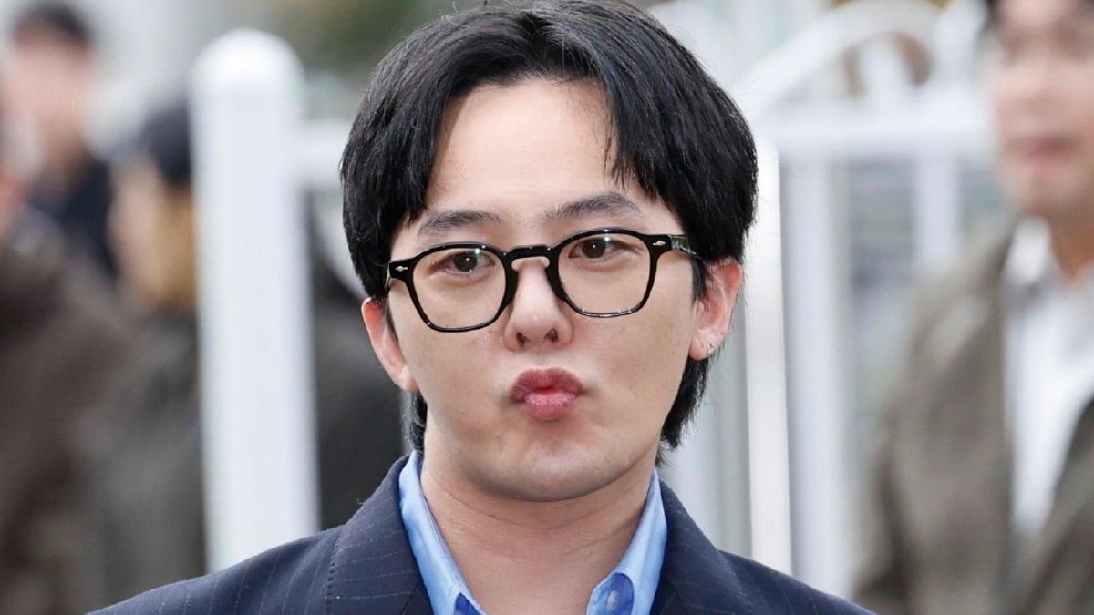 toc-g-dragon-12 Người đàn ông đeo kính đứng ngoài trời, mặc áo vest và áo sơ mi xanh, biểu cảm khuôn mặt đang chu môi.