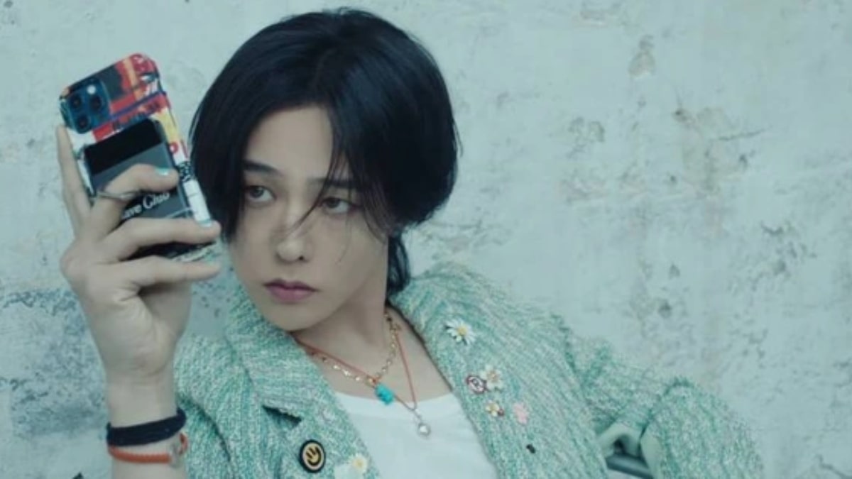 toc-g-dragon-13 Người cầm điện thoại, mặc áo khoác xanh nhạt, ngồi trước bức tường xám nhạt, nhìn về phía xa.