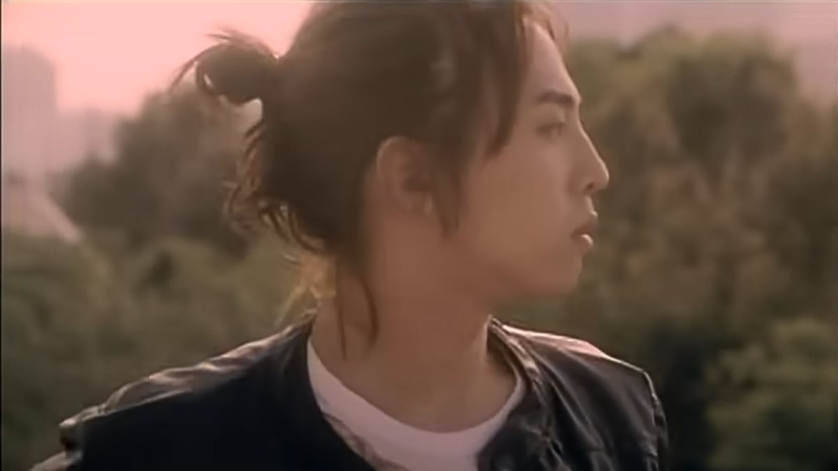 toc-g-dragon-14 Người mặc áo khoác da, tóc dài buộc, nhìn sang bên, nền là cảnh cây xanh mờ nhạt.