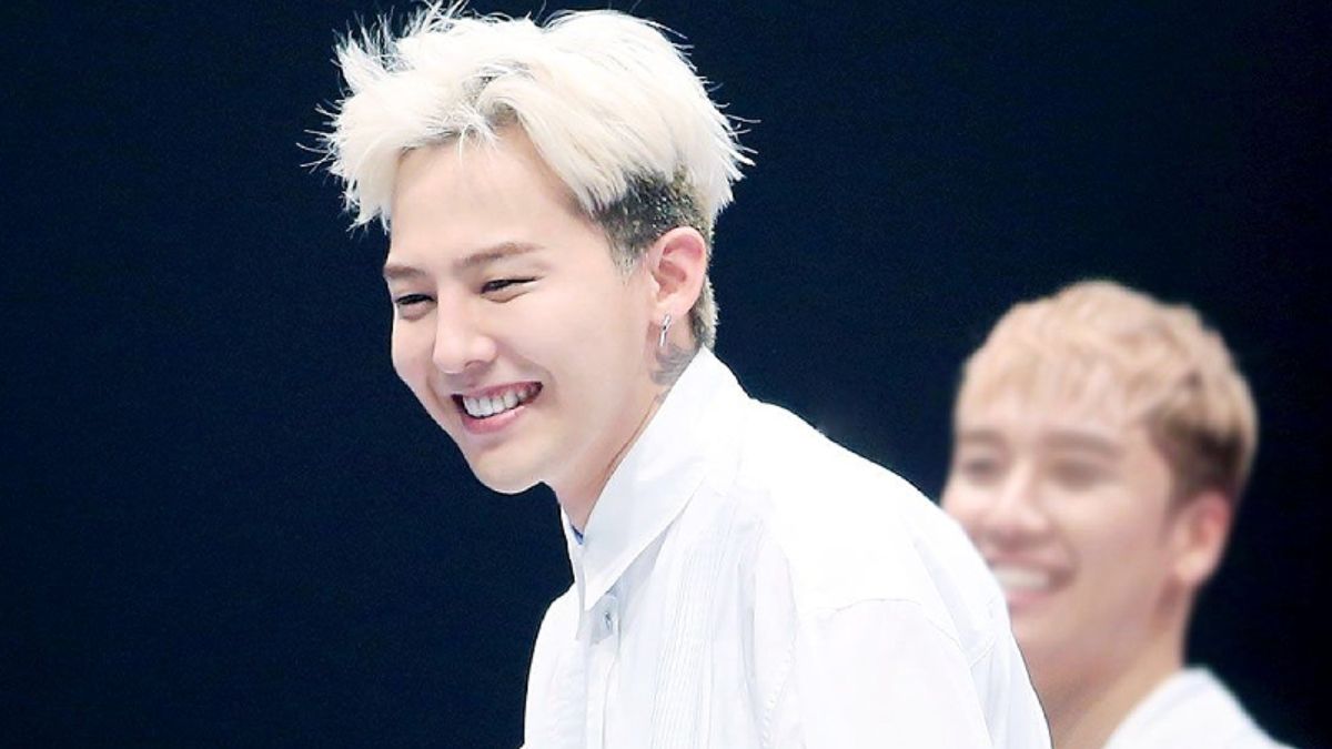 toc-g-dragon-15 Người đàn ông tóc vàng đang cười tươi, mặc áo trắng, nền tối, có một người khác mờ phía sau.