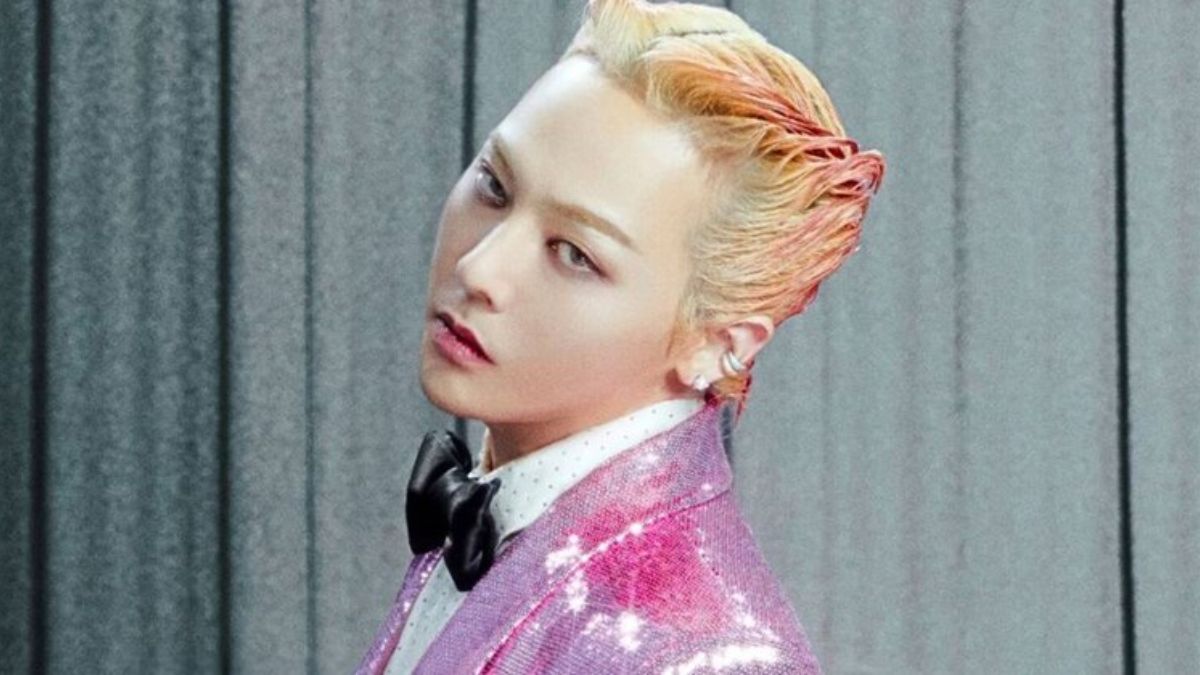 toc-g-dragon-17 Người mặc vest hồng lấp lánh, áo sơ mi trắng, nơ đen, tóc nhuộm sáng, đứng trước phông nền xám.