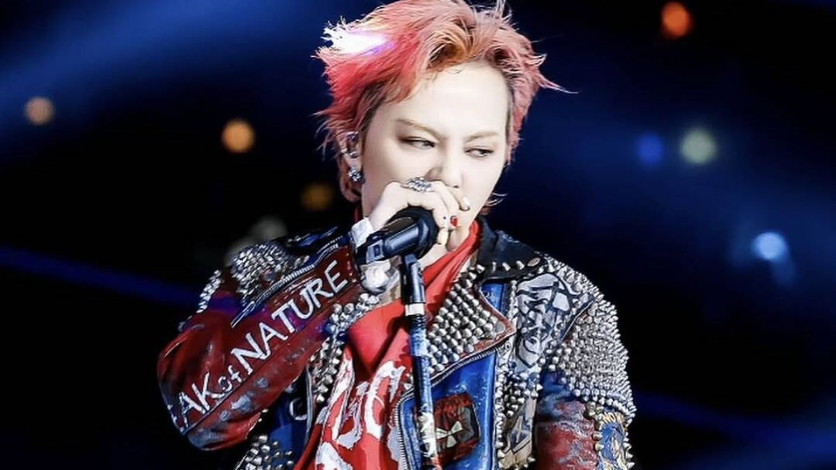 toc-g-dragon-19 Người trình diễn trên sân khấu, mặc áo khoác đính đinh và cầm micro, ánh sáng sân khấu rực rỡ phía sau.