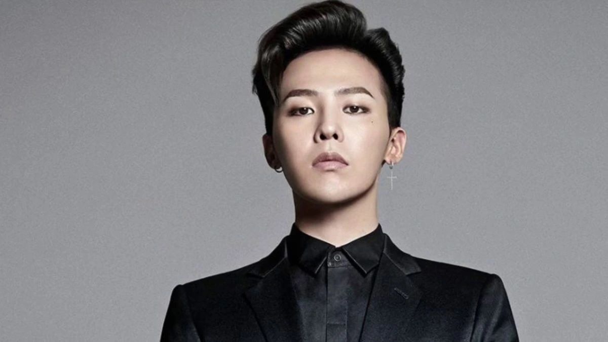 toc-g-dragon-2 Người mặc vest đen, đeo khuyên tai hình thánh giá, nhìn thẳng về phía trước trên nền xám.