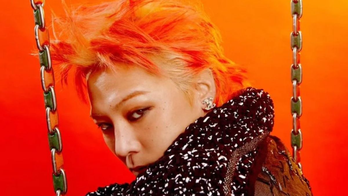 toc-g-dragon-20 Người có tóc màu cam nổi bật, đeo khuyên tai, nhìn về phía trước, nền màu cam, một phần dây xích bên cạnh.