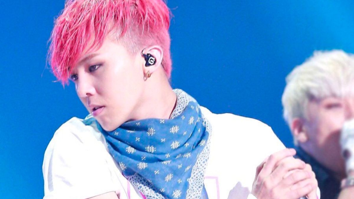 toc-g-dragon-21 Người có tóc hồng và khăn quàng cổ xanh đứng trên sân khấu với ánh sáng xanh phía sau.