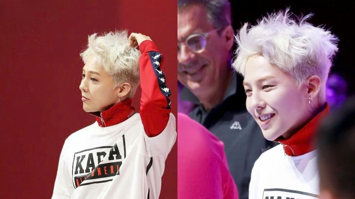 toc-g-dragon-24 Người tóc trắng mặc áo trắng, đứng trước nền đỏ, đang cười với một người khác trong khung cảnh sáng sủa.