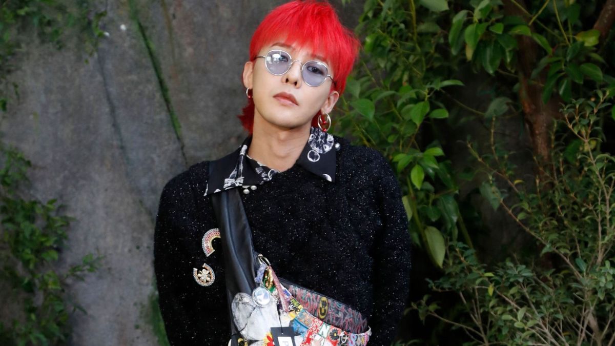 toc-g-dragon-25 Người tóc đỏ mặc áo đen, đeo kính tròn, đứng trước nền cây xanh và bức tường xám.
