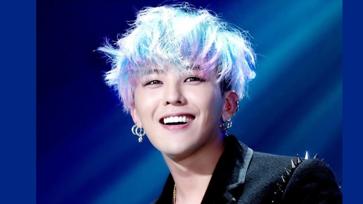 toc-g-dragon-26 Người cười rạng rỡ trên sân khấu, tóc nhuộm màu sáng, đeo hoa tai và mặc áo khoác đen.