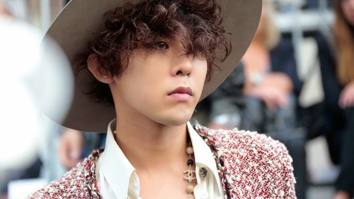 toc-g-dragon-9 Người đội mũ rộng vành, tóc xoăn, mặc áo khoác họa tiết, đứng trong không gian ngoài trời.