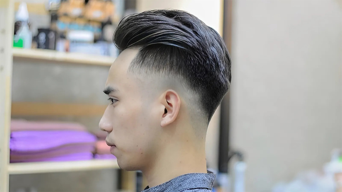 toc-gay-nhon-6 Người đàn ông với kiểu tóc undercut, phía sau là kệ gỗ chứa đồ vật trong một tiệm cắt tóc.