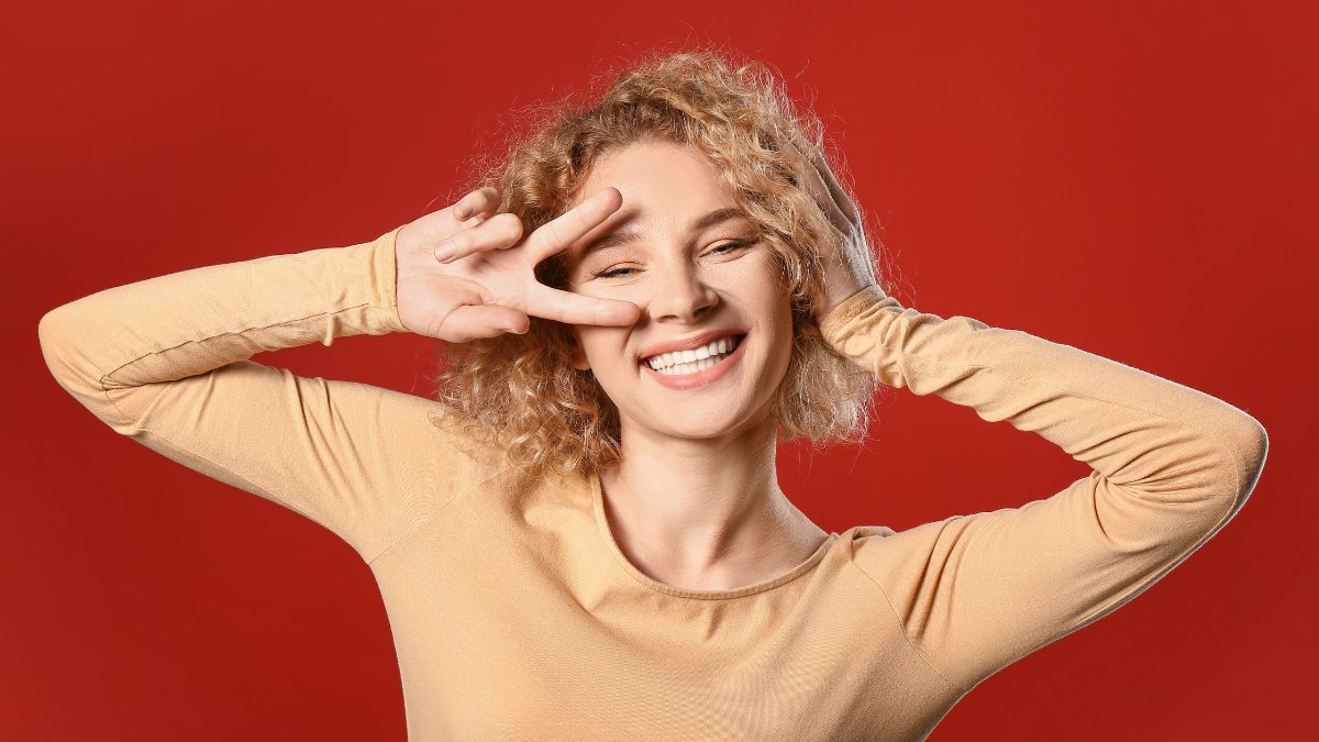 Người phụ nữ với mái tóc xoăn đang cười, tạo dáng vui vẻ với tay làm ký hiệu chữ V, nền đỏ.