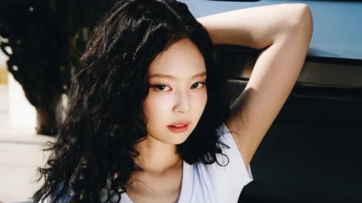 toc-jennie-5 Người phụ nữ mặc áo trắng, tóc xoăn đen dài, đang đứng ngoài trời với cánh tay giơ lên tựa vào xe.