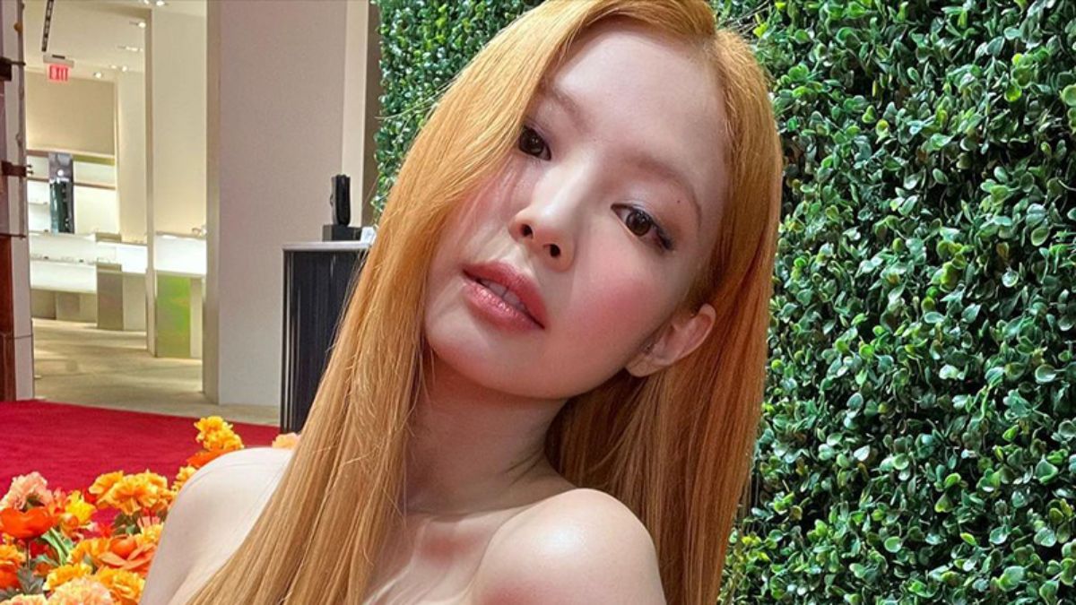 toc-jennie-6 Người phụ nữ tóc dài màu vàng đứng trước bức tường cây xanh trong một không gian nội thất.