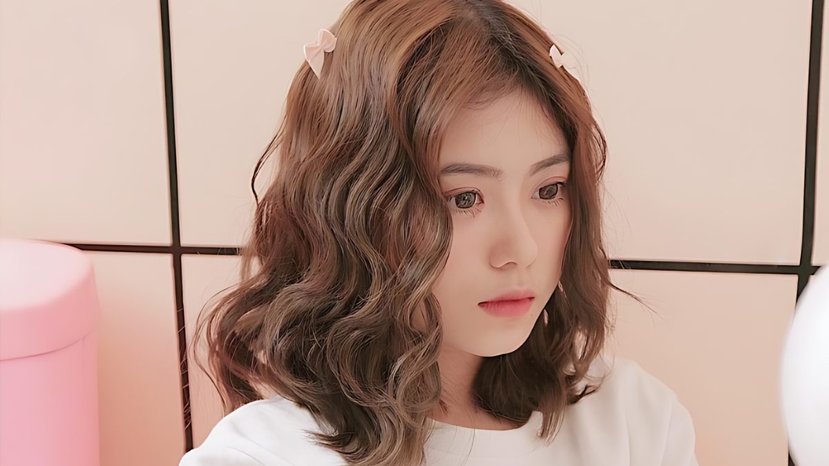 Người phụ nữ trẻ có tóc xoăn nâu, mặc áo trắng, ngồi trước nền tường màu hồng nhạt.