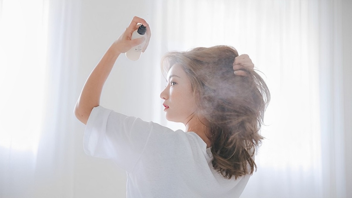 Người phụ nữ xịt sản phẩm lên tóc trong không gian sáng, cửa sổ có rèm trắng nhạt phía sau.