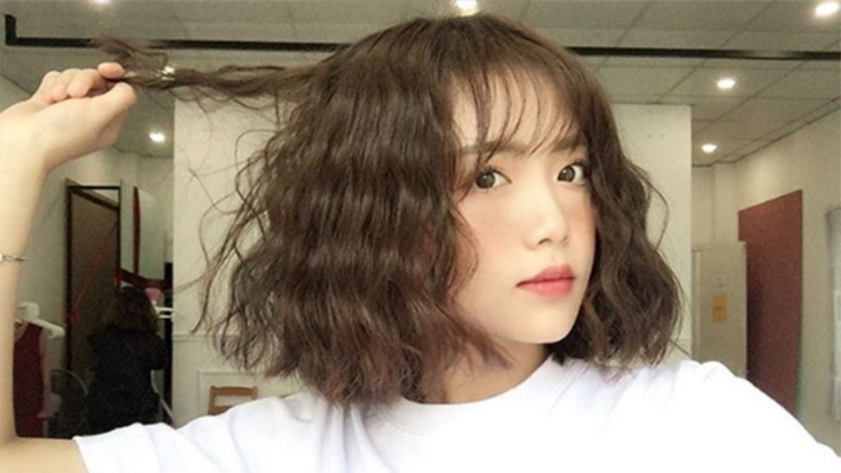 Người phụ nữ tóc ngắn xoăn, mặc áo trắng, đang tạo dáng trong phòng sáng.