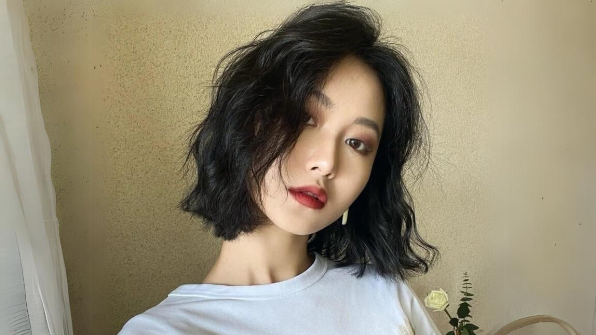 Người phụ nữ với tóc ngắn xoăn, mặc áo trắng, chụp ảnh trước nền tường màu be.