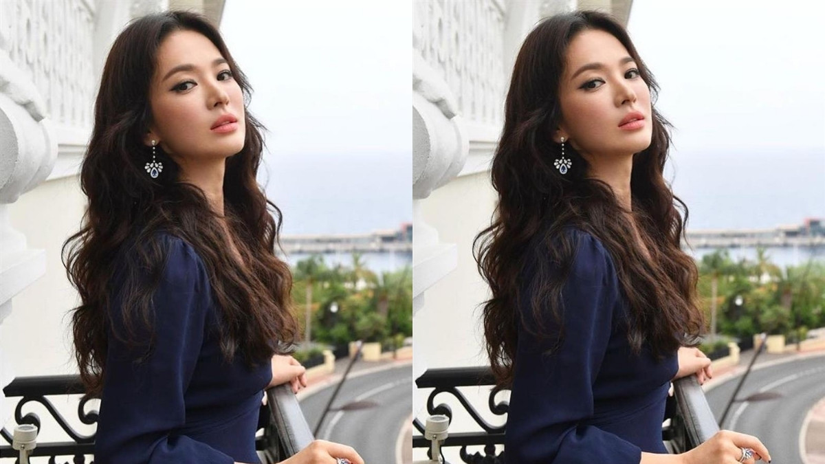 toc-song-hye-kyo-4 Người phụ nữ đứng tựa lan can, mặc áo xanh đậm, tóc dài xoăn, phông nền là phong cảnh đường phố gần biển.