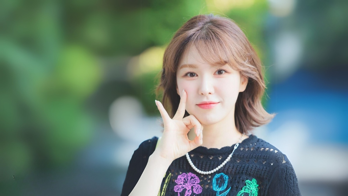 toc-wendy-4 Người phụ nữ trẻ đứng ngoài trời, mặc áo đen có họa tiết hoa, cười tươi và đặt tay lên má.