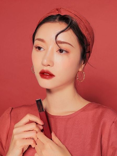 Người phụ nữ cầm thỏi son, mặc áo và khăn cài đầu cùng màu, nền màu đỏ.