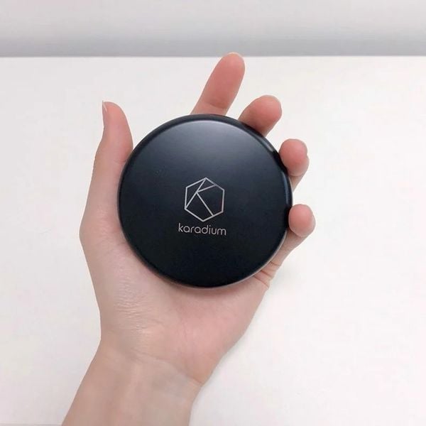 Một tay cầm hộp phấn trang điểm màu đen, trên bề mặt có logo và chữ "karadium".