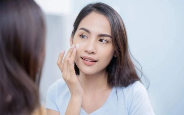 Người phụ nữ thoa kem dưỡng da lên mặt, mặc áo màu xanh nhạt, đứng trước gương trong phòng sáng.