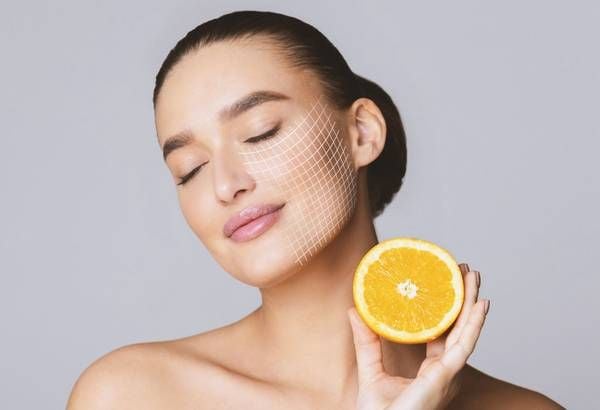Người phụ nữ cầm nửa quả cam, mắt nhắm và có lưới đồ họa trên mặt, nền màu xám nhạt.