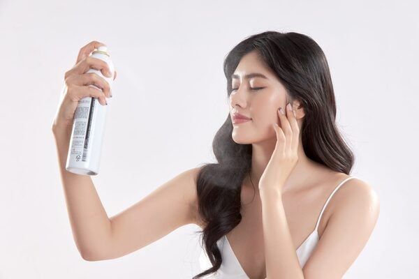Người phụ nữ đang sử dụng bình xịt dưỡng da, mắt nhắm, biểu cảm thư giãn trên nền trắng.